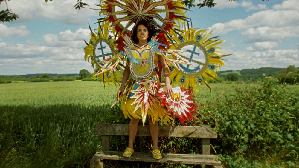 Artist Rhea STorr, a mixed race woman wears an elaborate carnival costume decorated with feathers and circular structures around her head. She stands on a bench, looking straight at the camera against a vast green field and blue sky. 