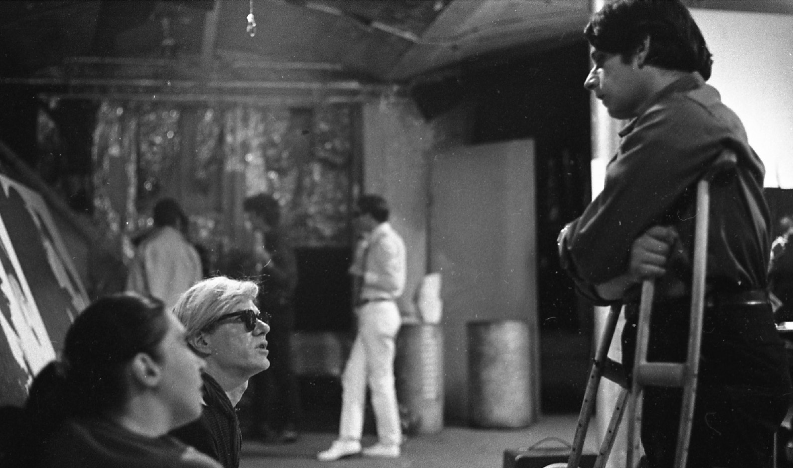 a black and white image, on the left hand side Andy Warhol with white hair and dark glasses looks up at Stephen Dwoskin who stands resting on a pair of crutches looking down at him