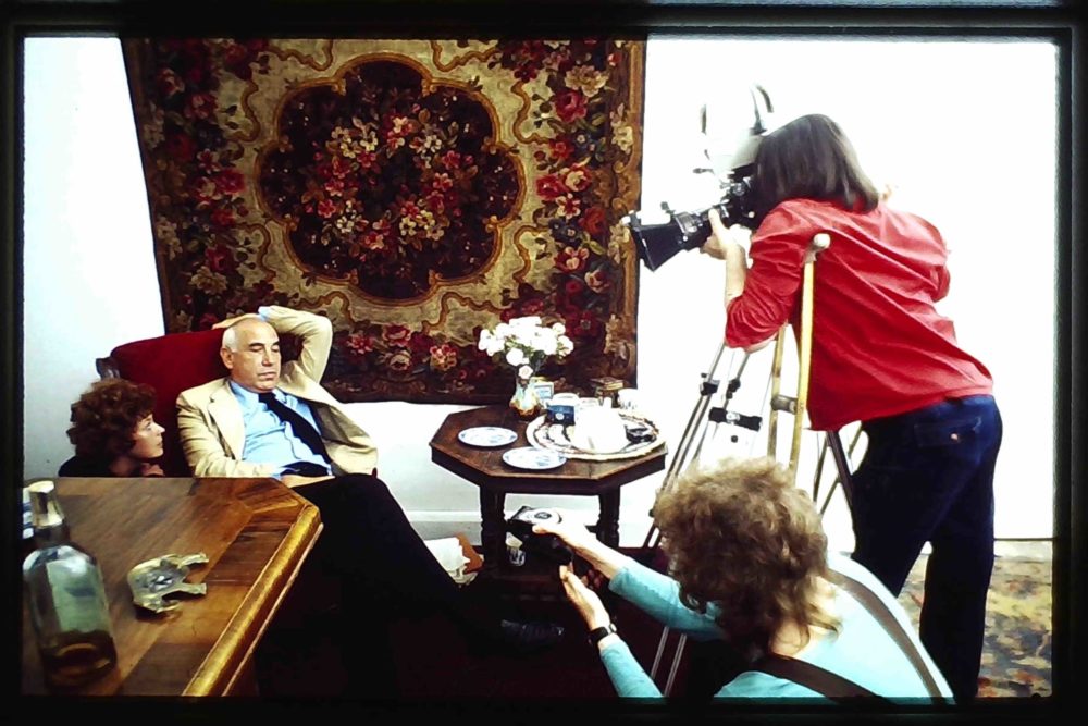 a person a crutch and another person with a recorder are filming man slouched in an armchair between two tables against a wall carpet. A woman perches on the armchair.