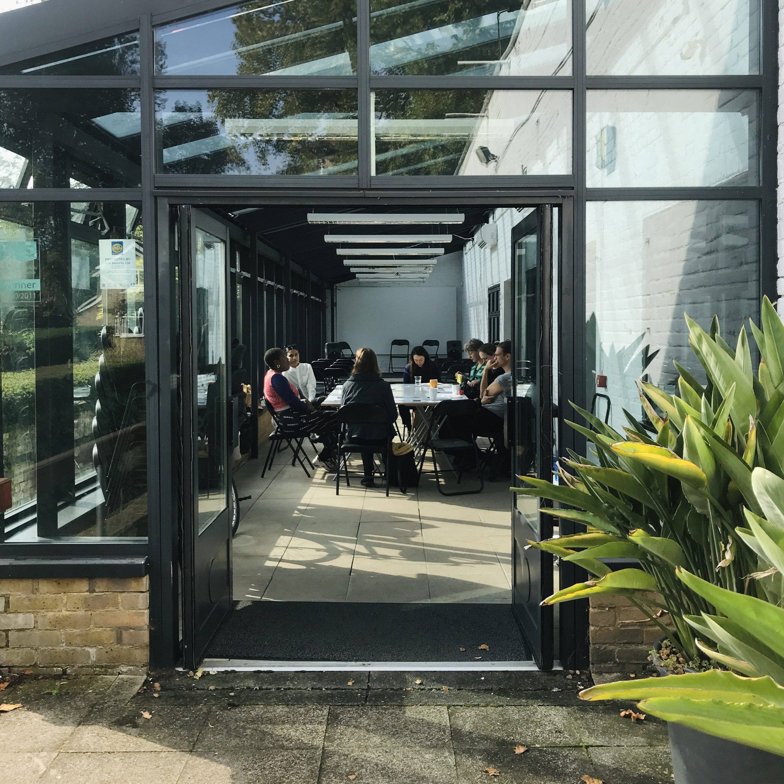 image of the conservatory at LUX from outside looking through an open door. A group of people sit in intense conversation.