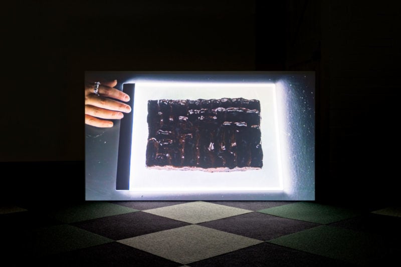 Installation of a screen standing on a checkered floor. On a screen is an image of a rectangular dark brown slab of ceramic piece with rough surface and a hand touching the power button of the lightbox on the top left corner.