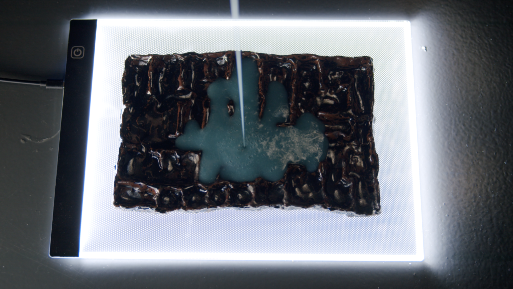 A blue opaque liquid viscously drizzles from above and fills a lumpy rectangular ceramic that sits on a glowing lightbox