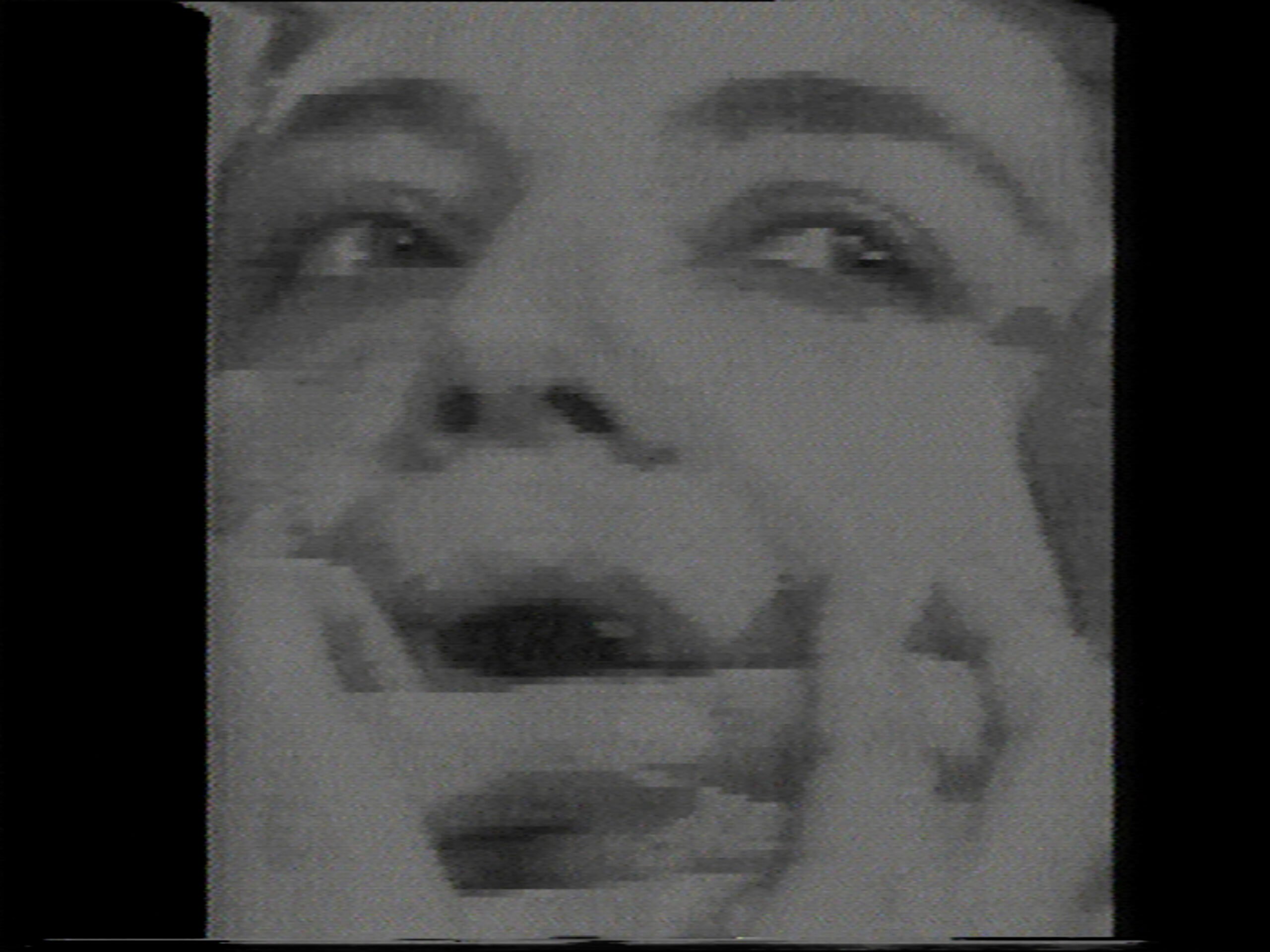 Still from Mona Hatoum’s So much I want to say. A low res black and white close up of a face whose mouth is covered by a hand, gagging. 
