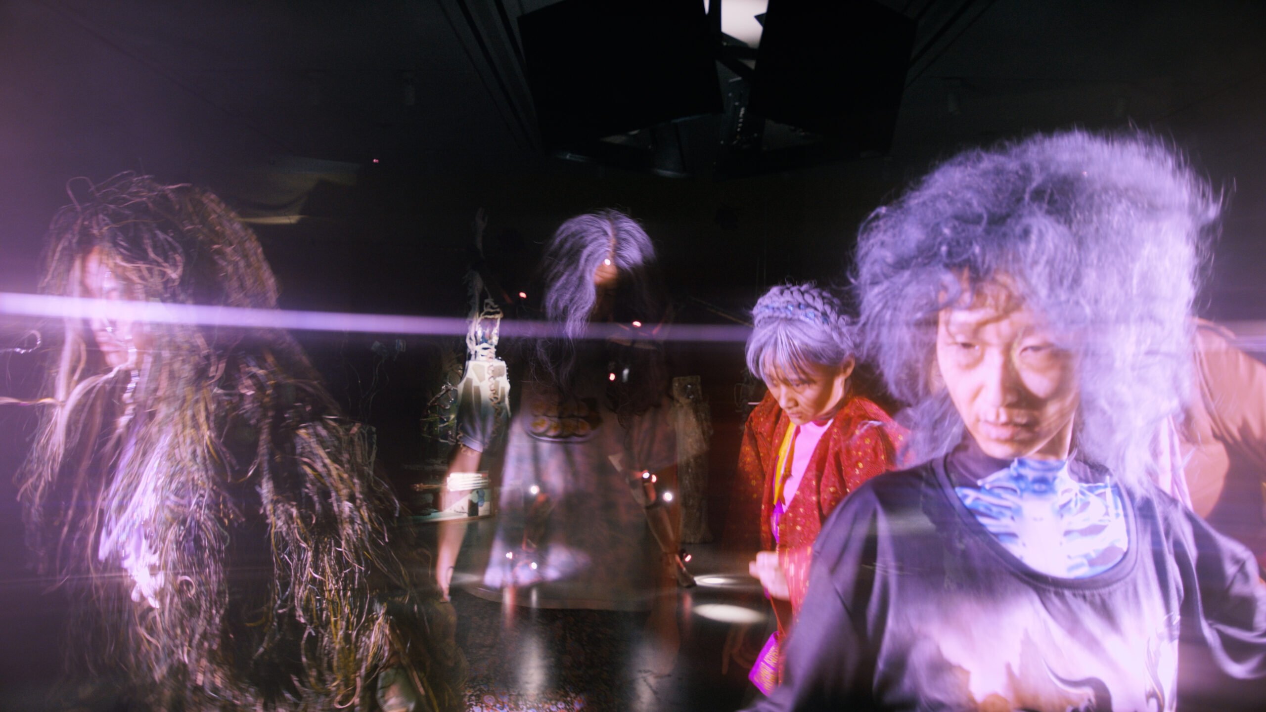A dark empty gallery is overlaid with a ghostly images of four performers in old age makesup and wigs with blank stares.