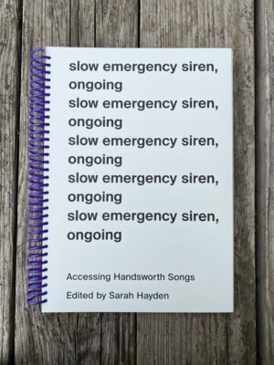 a white book with a large purple spiral binding placed on a wooden surface. On the cover is the title slow emergency siren, ongoing written five times and below the title is subtitles that read accessing handsworth song edited by Sarah Hayden