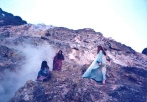 Three women in long dresses on a volcanic mountain where volcanic fumes billowing out from the crater.