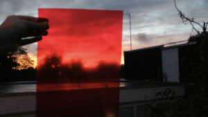 A hand holding a red translucent rectangle that filters the bright orange sunset emerging through cloudy sky into a blurry red tinted trees and sky
