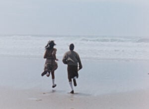 The back of two students in their school uniforms - skirts, cardigans and white socks - running towards the ocean waves crashing on sandy, sun bleached shore.
