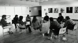 a black and white image of a group of people sitting on chairs in deep discussion in a gallery space