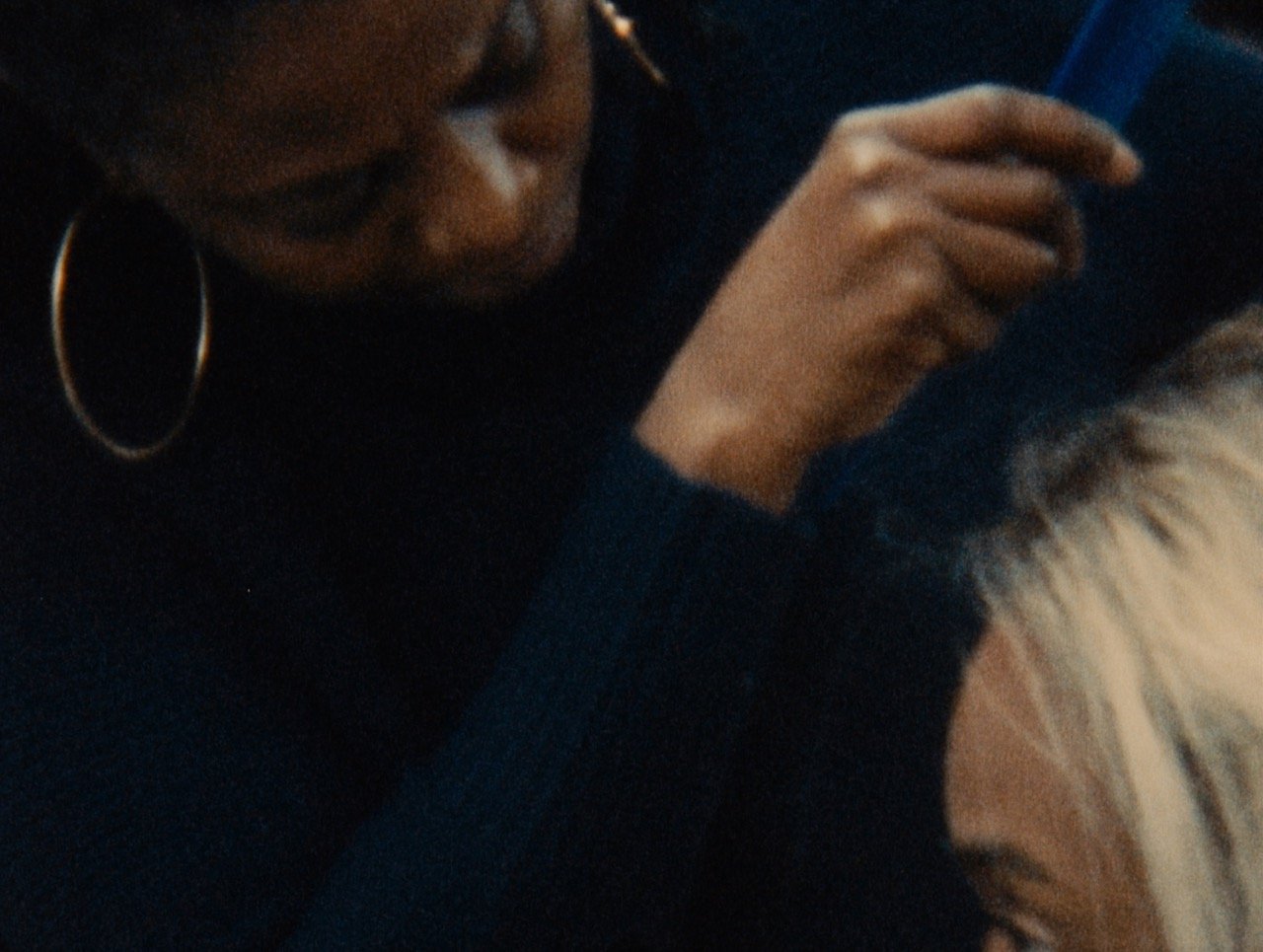 A close look at a black person with large hoop earrings in a navy turtleneck looking closely at another person and gesturing to comb their grey hair that is barely visible in the frame