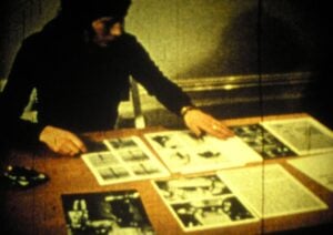 A person in a black turtleneck examines paper documents with words and images laid out on a wooden table.