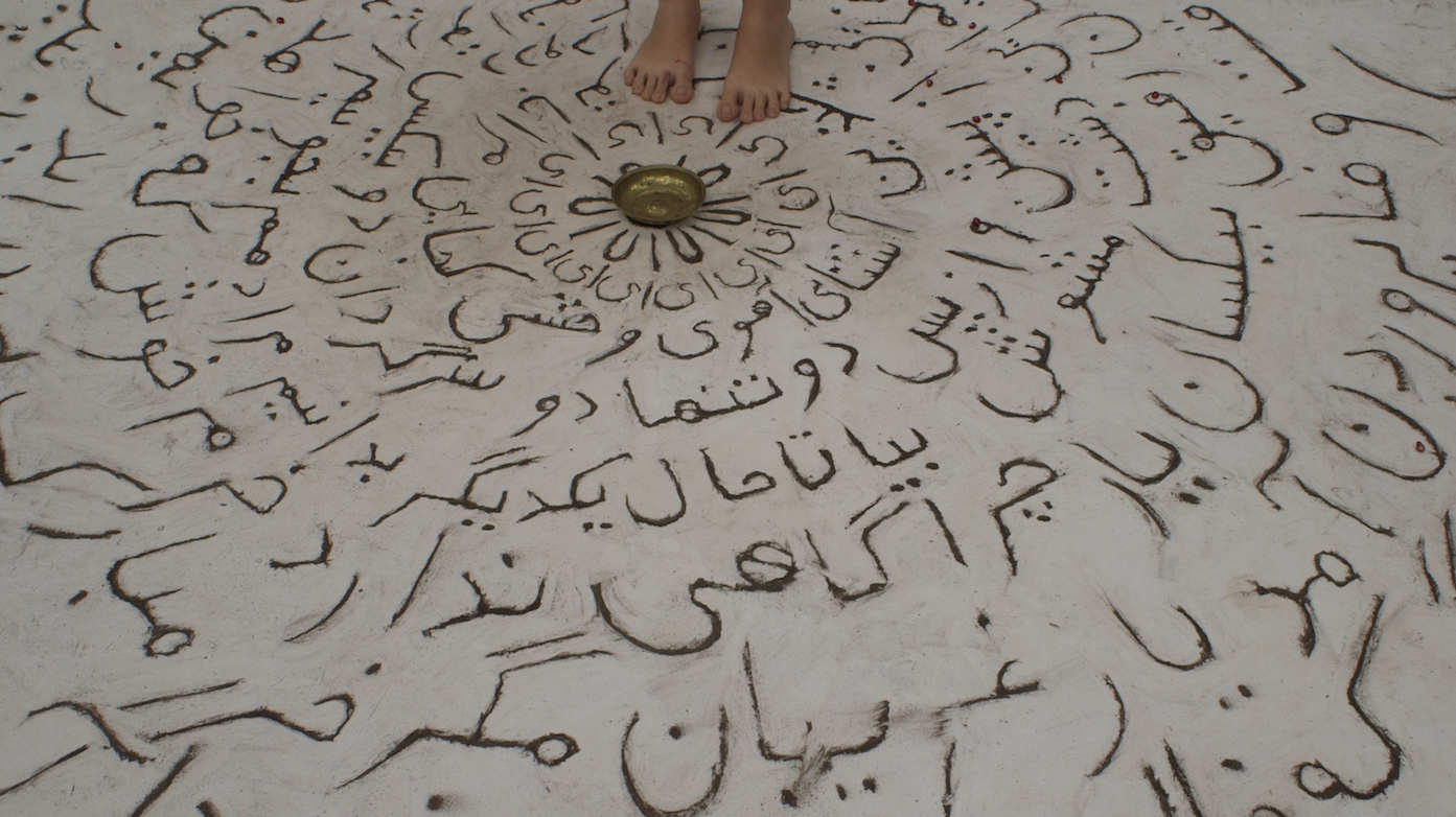 Persian alphabets are hand-written in earth on a white floor, forming concentric circles that surround a metal bowl and a pair of bare feet. The circular shapes create a sense of depth and perspective, drawing the viewer's eye toward the center of the image