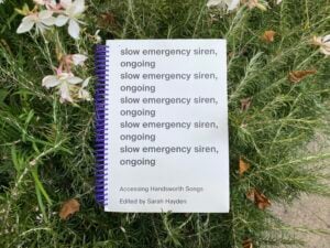 We see a white book with a large purple spiral binding lie on a bed of rosemary and a few white flowers. The cover has its title in black that reads “slow emergency sire, ongoing” written five times.