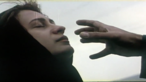 A woman in a hijab with her head slightly tilted back, eyes closed, and a serene expression. A hand stretches into the frame, as if reaching towards the woman's face