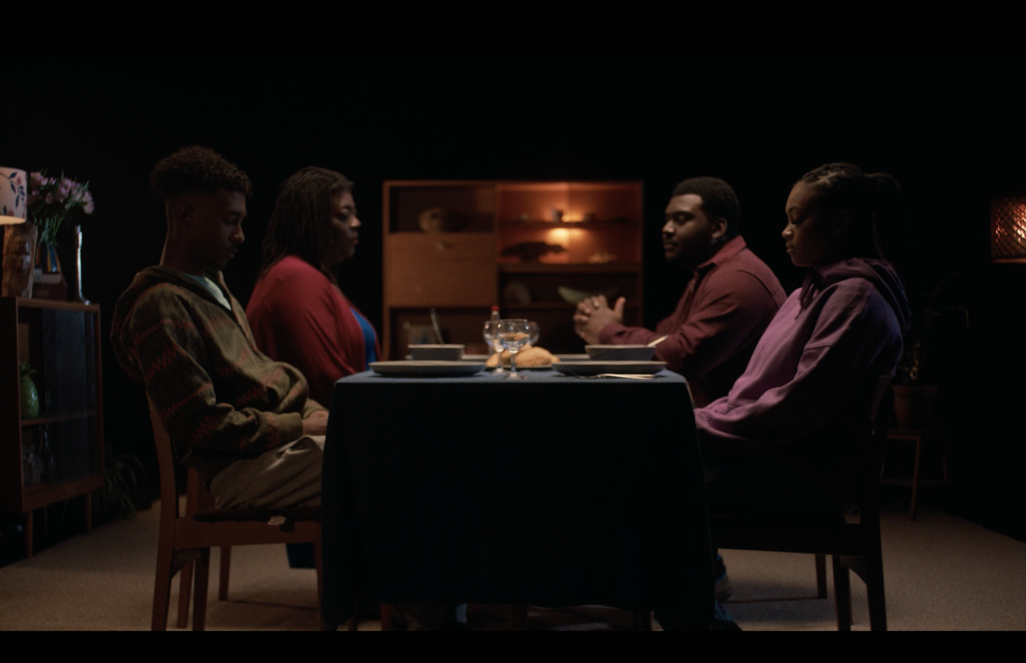 In a sound stage with black walls, a family of four individuals with dark skin is seated around a table. They wear solemn expressions, illuminated by soft, dim lighting. The warm glow of lamp lights highlights the wooden furniture in the background