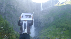 A person holding a piece of paper with a word ‘STOPP’ printed in black and white, obscuring the head of the person. Behind the person is a mountain range with two waterfalls