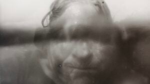 In this black and white image, a face is partially submerged in water, captured by a partially submerged camera. The water acts as a magnifying lens, distorting and enlarging the lower half of the face. Above the waterline, the forehead emerges, illuminated by a glow from the sunlight