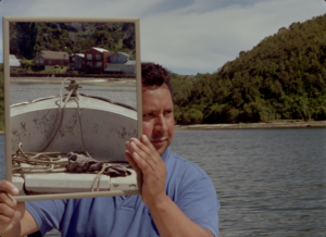 a still image from Double Ghosts by George Clark, a man holds up a mirror and looks to the side it reflects the front of a small boat and shoreline.