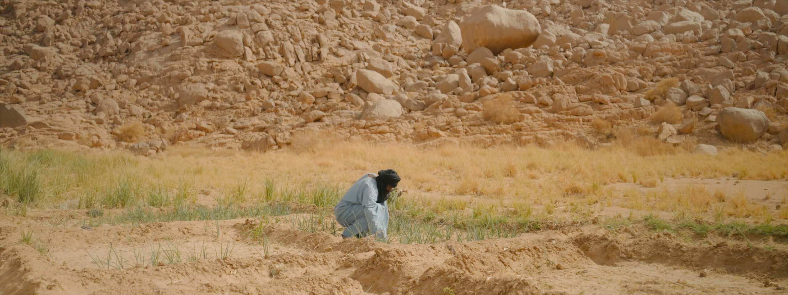 A person wearing a grey outfit and a black turban squats in the middle of a sandy ground with patches of green and yellow grass. The background is a steep, rugged rocky slope