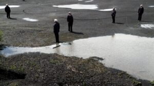 Five people in matching dark uniforms stand apart from each other on a moss-covered ground, surrounded by muddy puddles. Each person faces a different direction