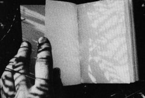 a film still from Guy Sherwin's 'Messages'. A black and white image of a hand holding open a book with blank pages, with the shadows of branches falling across it
