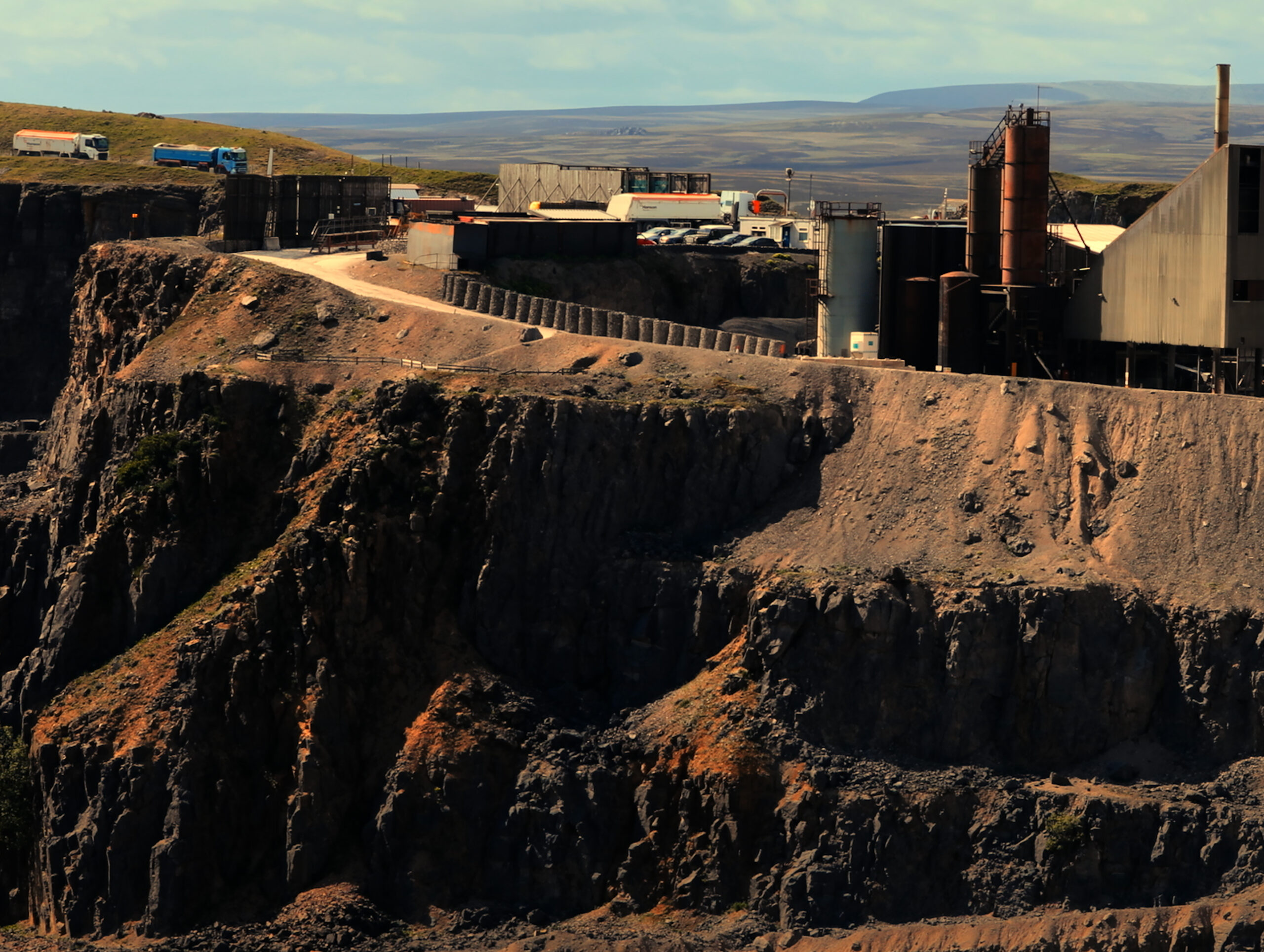 Atop a rugged cliff is an industrial site with cylindrical metal structures, accompanied by a parking lot filled with containers and cars. A couple of trucks are making their way into the site