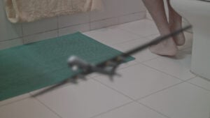 Image of a white tiled bathroom floor with a green bath mat. Bare legs and feet can be seen on the centre of the image. A silver model plane is in the centre of the frame, out of focus.