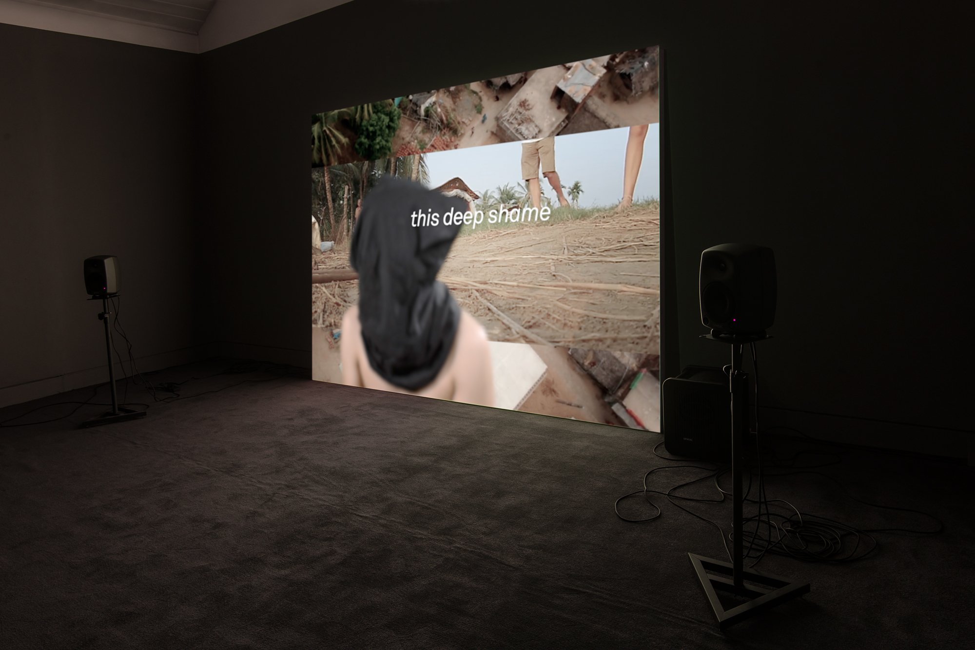 An exhibition installation featuring a large projection screen at the centre, flanked by speakers on each side. The screen displays a collage of digital images, in the foreground is an image of a person wearing a cloak over the head and bared torso. The person is in soft focus and is overlaid with text that reads: "this deep shame."
