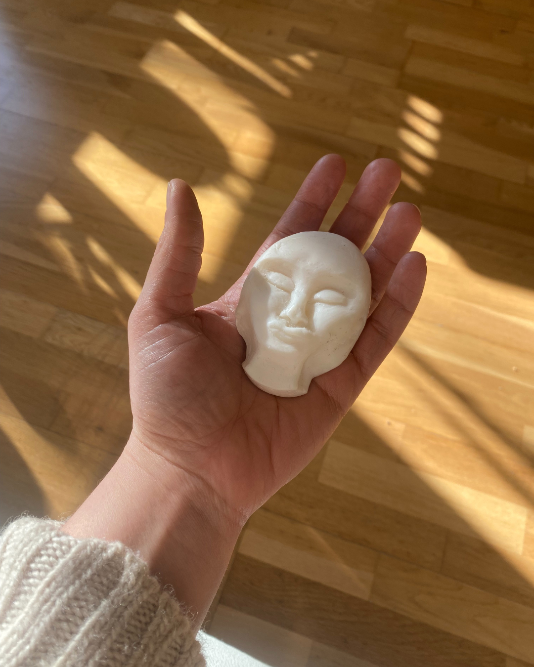 A bar of soap carved into the shape of a face rests on a palm on a wooden floor, illuminated by sunlight streaming through a window.