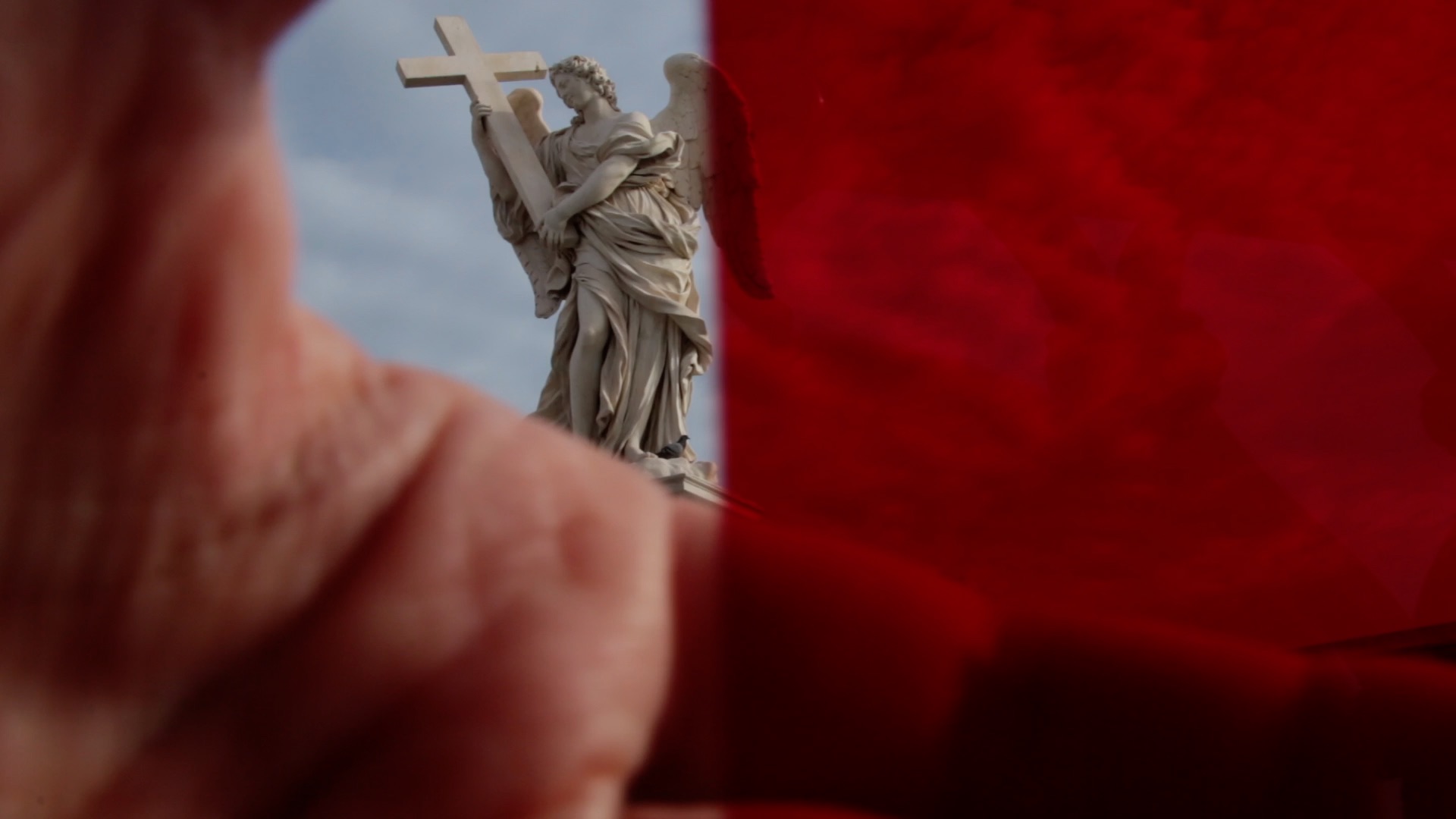 Image of a statue of an angel with wings holding a cross in the background and in the foreground, a hand slightly covering the lens on the left side of the frame and the entire right side of the frame is tinted red.