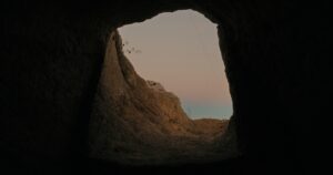 Image from inside a cave looking up and out to the walls of the cave and the sky that is a gradient of grey, to pink, to blue. The grey and pink being clouds, and the blue being the sky.