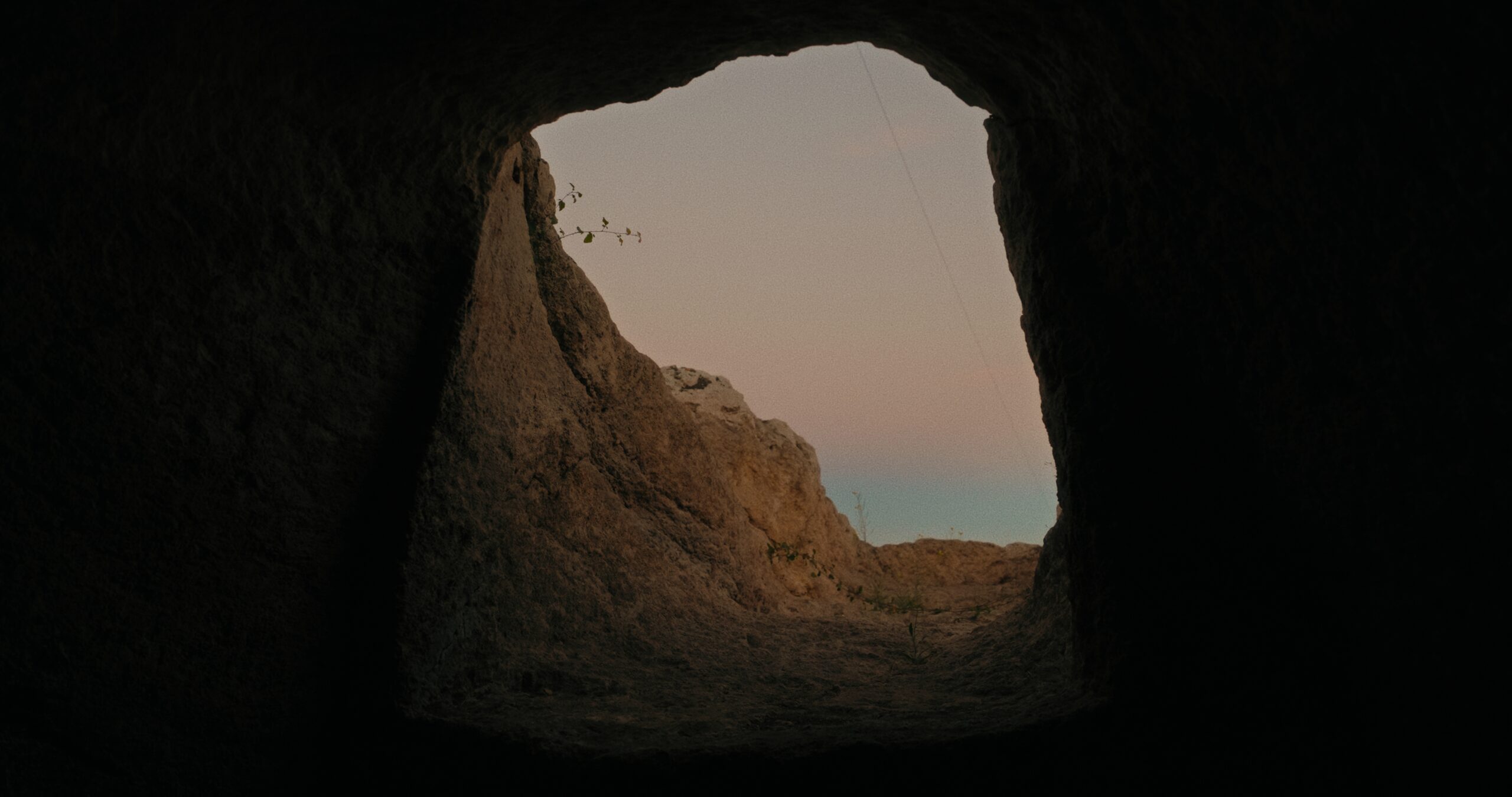 Image from inside a cave looking up and out to the walls of the cave and the sky that is a gradient of grey, to pink, to blue. The grey and pink being clouds, and the blue being the sky.