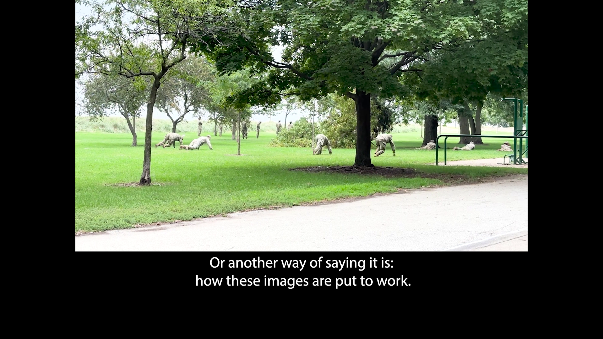 Soldiers lower to the ground, doing a training exercise in a green space in a park, framed by a black border. Beneath the image there’s a caption that reads: “Or another way of saying it is: how these images are put to work.”