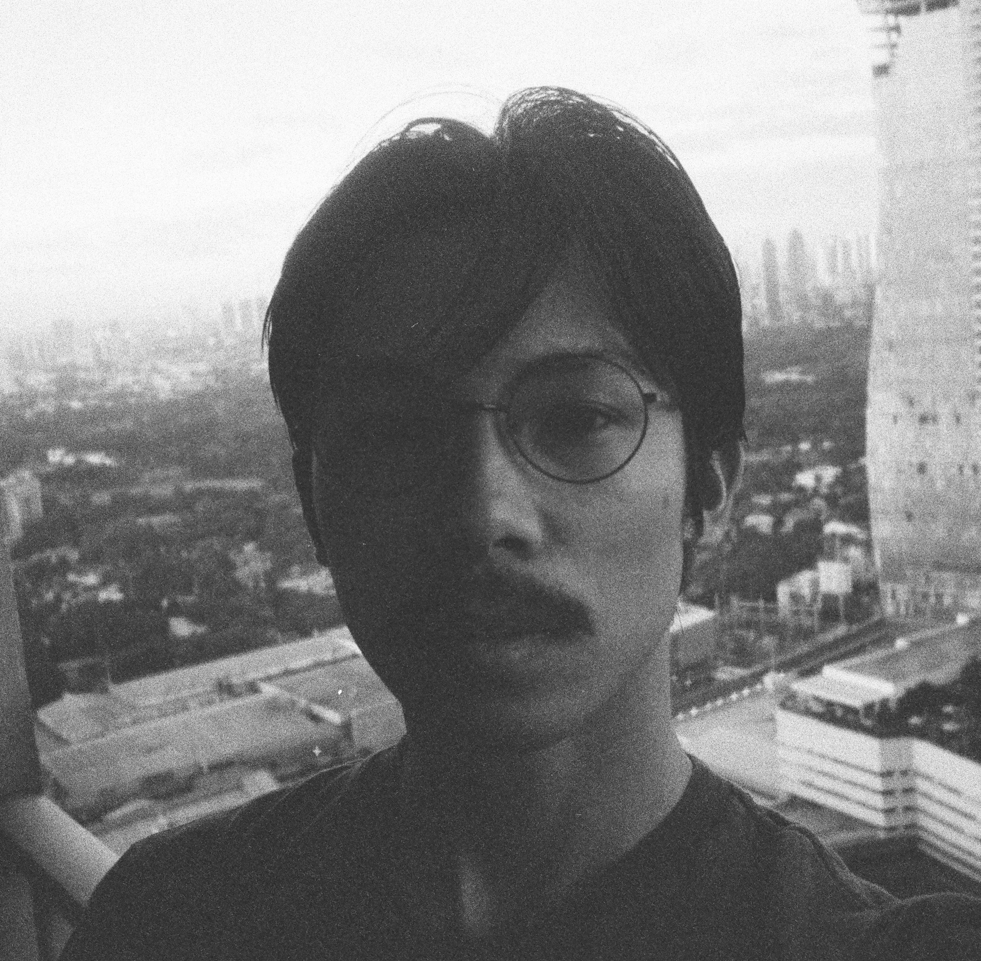 Black and white photograph of filmmaker Miko Revereza standing on what appears to be a balcony overlooking a cityscape. He is wearing round glasses and has a moustache, with his face partially in shadow. Tall buildings and a skyline can be seen in the background.