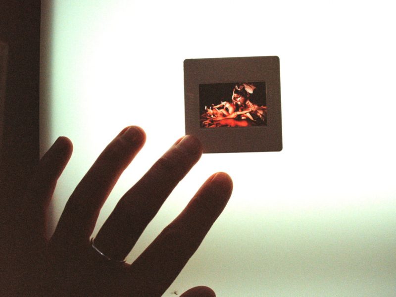 A colour positive photography slide lies flat on a light box. The frame captures several naked bodies mingled and piled on top of each other. A hand from the left corner touches the slide with a tip of its finger.