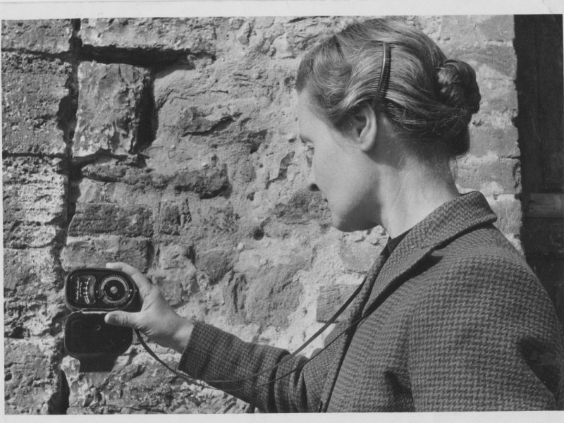 black and white image of filmmaker Margaret Tait, wearing a formal jacket, she is looking away from the camera at a light meter and is framed against a brick wall