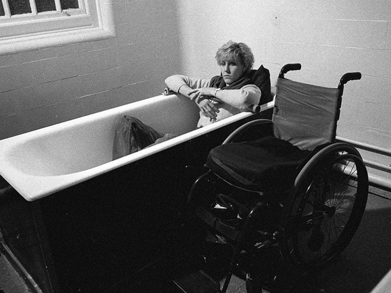 Ellen Stohl, a white woman with a short cut is sitting in a bathtub. A wheelchair stands next to her.