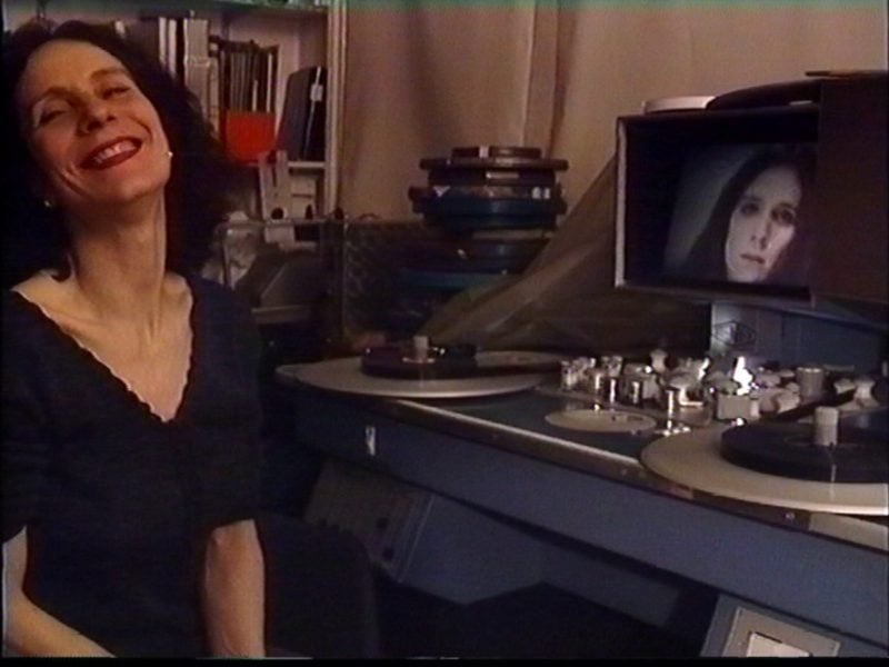 A white woman in a black dress sits next to a steenbeck showing a big happy smile to the camera. On the steenback’s monitor is showing a younger portrait of the woman.