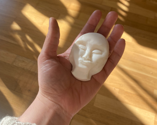 A bar of soap carved into the shape of a face rests on a palm on a wooden floor, illuminated by sunlight streaming through a window.