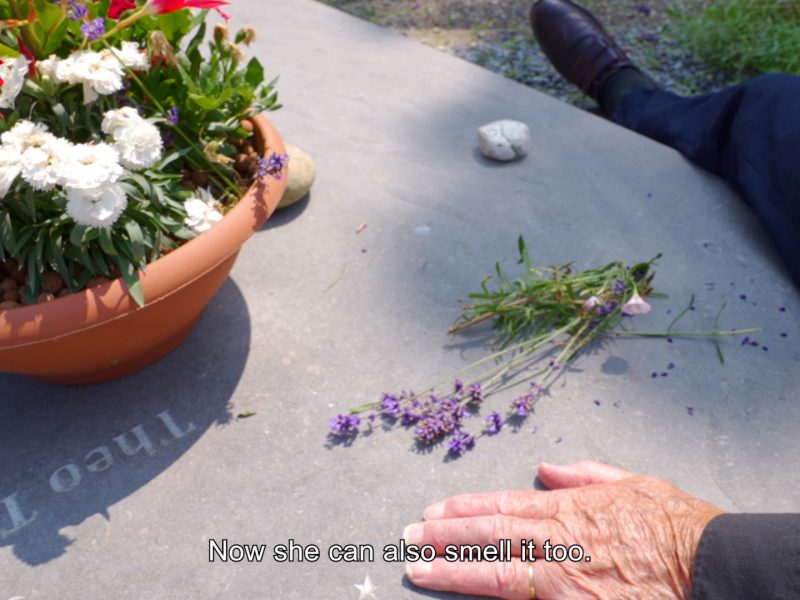 On a gravestone are flowers in a pot and a few lavender flowers laid flat. A hand of a whie elderly person touches the gravestone next to flowers. The caption reads “Now she can also smell it too”.