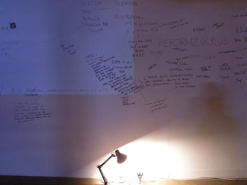 Dimly lit room with a white wall covered in hand-written mind maps. A blue rectangular projection casts a soft glow, contrasting with the bright spotlight of a table lamp on the floor