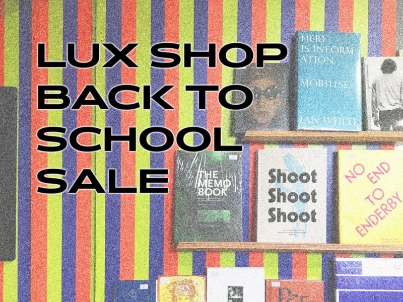 A black caption with white outlines that reads 'LUX Shop Back to School Sale' against a faded, grainy image featuring colorful stripes and three tiers of book displays