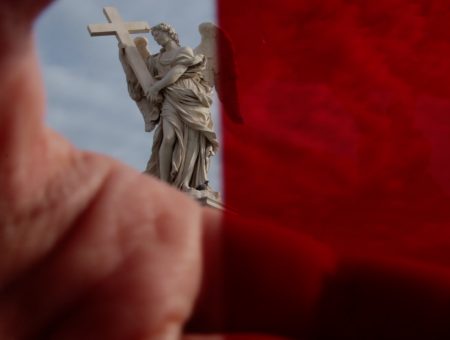 Image of a statue of an angel with wings holding a cross in the background and in the foreground, a hand slightly covering the lens on the left side of the frame and the entire right side of the frame is tinted red.