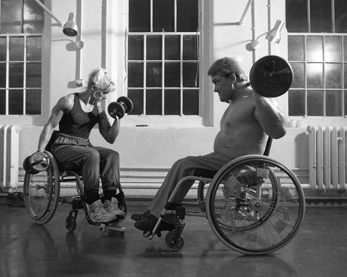 A white woman and white man in wheelchairs weightlifting in a well-lit indoor space.