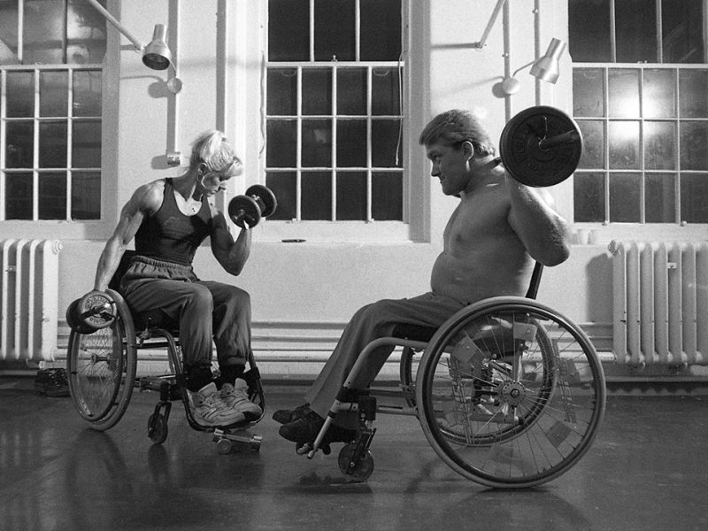 A white woman and white man in wheelchairs weightlifting in a well-lit indoor space.