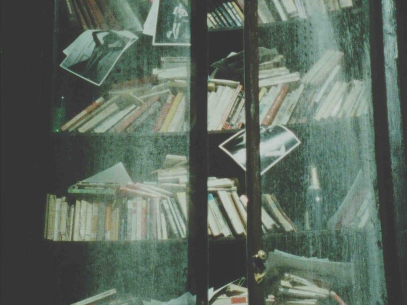 A bookcase with dirty glass doors is fulled with books and photographs tucked underneath the books to make them visible.