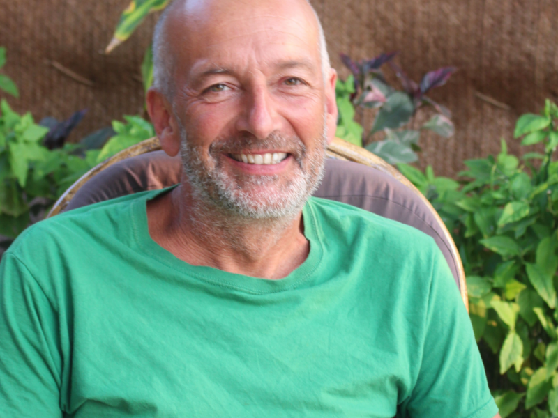 portrait of George Barber, a middle aged white man with a bald head and grey beard wearing a green t-shirt, smiling and sitting in front of some plants