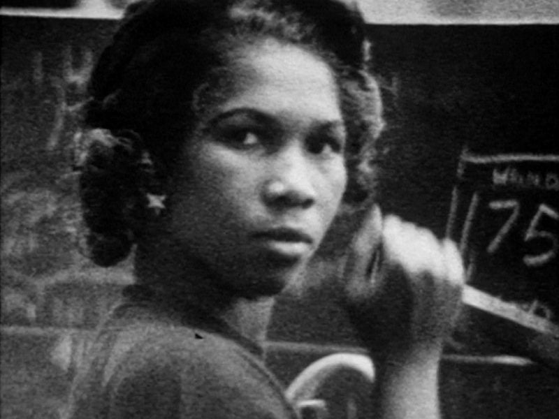 A grainy black and white image captures a black woman who turns to look straight into the camera while her hand is raised as if she is holding something
