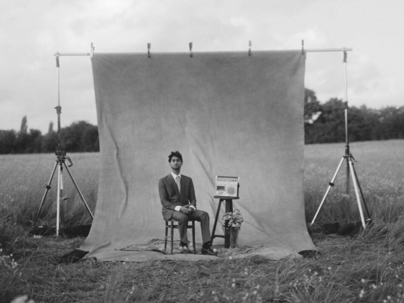 A person in a suit poses next to a cassette tape machine for a photoshoot. The backdrop is hung from c-stands in the middle of a field