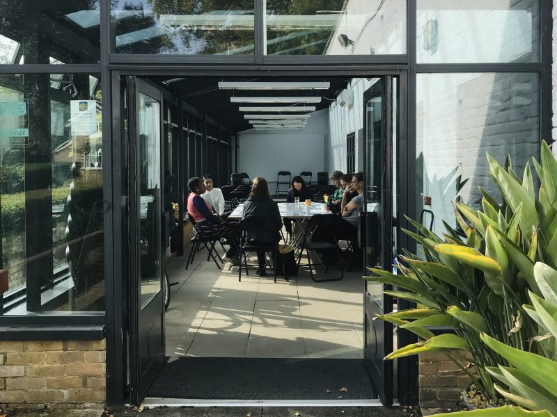 image of the conservatory at LUX from outside looking through an open door. A group of people sit in intense conversation.